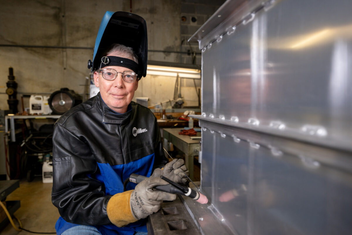 a man in a pushed back protective welding helmet poses so his face is visible, and he is in an industrial looking shop