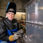a man in a pushed back protective welding helmet poses so his face is visible, and he is in an industrial looking shop