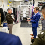 Two men, one in a suit and one in an army officer coat, talk to each other in a research lab