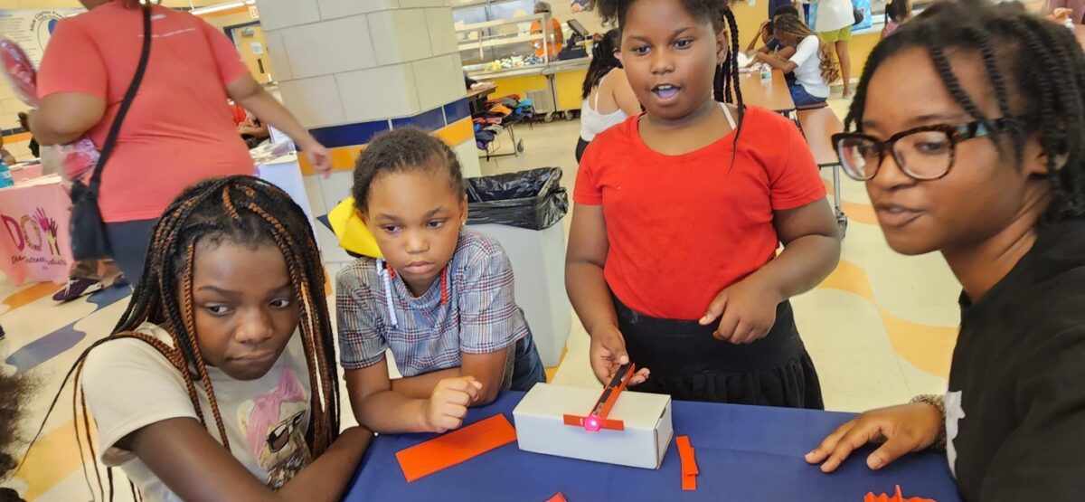three students sit around a table with a teacher, one is holding a laser pointer