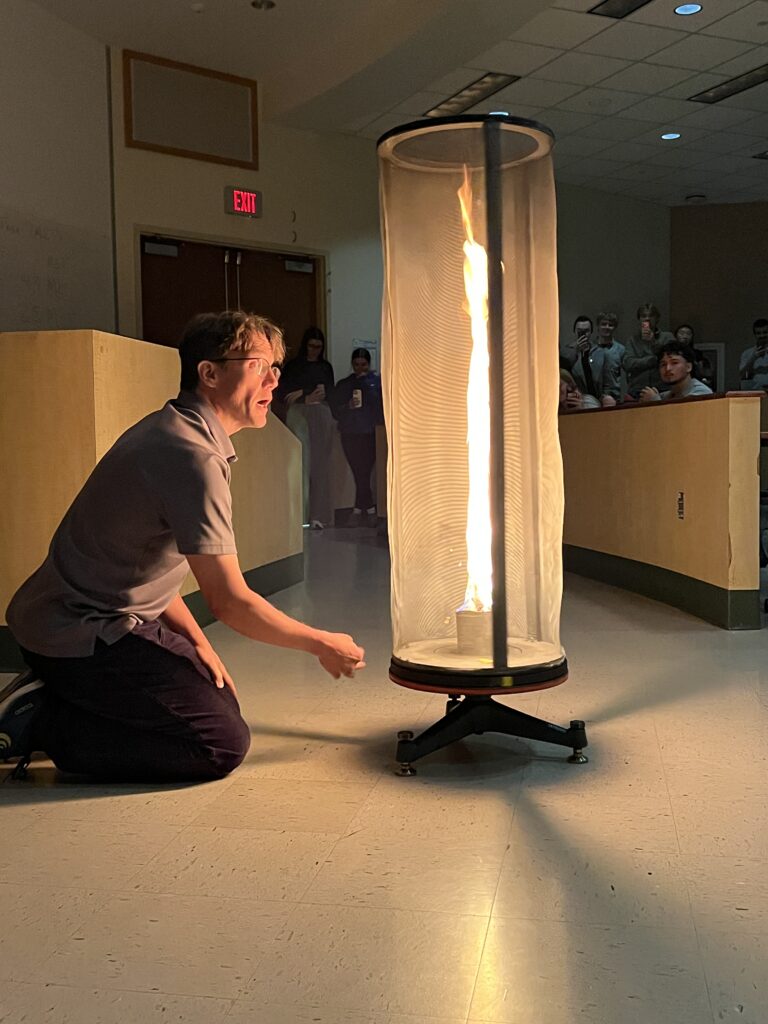 a man kneels down next to a tall mesh cylinder with a vertical column of fire inside it
