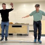 two people stand and balance on either side of a plank with a fulcrum under it. both their arms are out like they are trying not to fall.