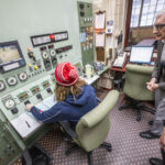 a person with their back to the camera adjust controls on a large control panel while the WI governor looks on